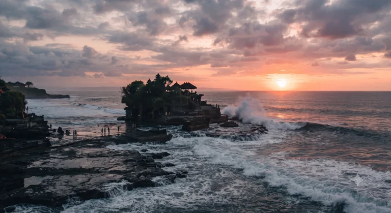 Balinese sea temple on a rocky islet at sunset with waves and visitors on the shore.