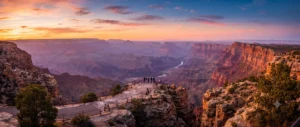 Sweeping sunset panorama of the Grand Canyon South Rim with visitors standing along the natural stone viewing platform.